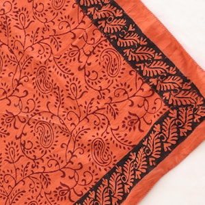May include: A square textile featuring a vibrant orange base color with a dark brown printed pattern. The design includes intricate paisley motifs and scrolling vines across the center, framed by a wide border of dense, repeating leaf shapes.