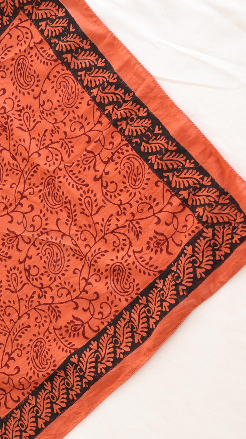 May include: A square textile featuring a vibrant orange base color with a dark brown printed pattern. The design includes intricate paisley motifs and scrolling vines across the center, framed by a wide border of dense, repeating leaf shapes.