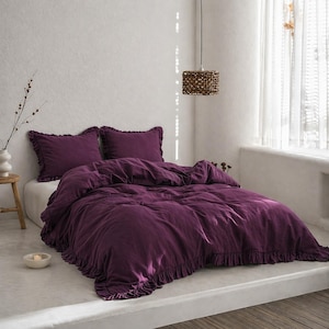 May include: A bed dressed in deep purple bedding, including a duvet cover and two pillowcases, all with a ruffled trim. The room has a neutral colour scheme, a woven pendant light, and a small side table.