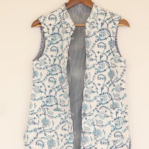 May include: A sleeveless, open vest with a floral pattern in shades of blue on a white background. The vest is hanging on a wooden hanger. The interior lining features vertical navy blue and white stripes.