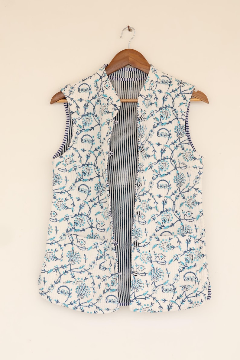 May include: A sleeveless, open vest with a floral pattern in shades of blue on a white background. The vest is hanging on a wooden hanger. The interior lining features vertical navy blue and white stripes.