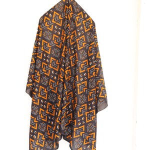 May include: A dark scarf with a geometric pattern in orange, white, and black hangs against a white background. The scarf's design features angular shapes and small dotted details, creating a striking visual contrast.