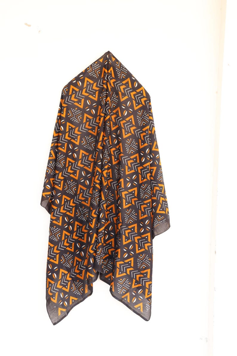 May include: A dark scarf with a geometric pattern in orange, white, and black hangs against a white background. The scarf's design features angular shapes and small dotted details, creating a striking visual contrast.
