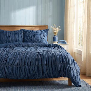 May include: A navy blue bedspread and pillow set with a gathered design. The bed is made up with a wooden headboard, a blue rug, and a blue vase with dried flowers. The room has light blue walls and beige curtains.
