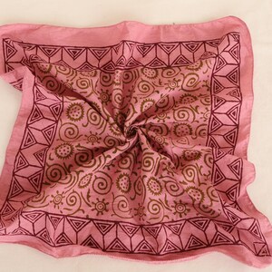 May include: A square pink bandana featuring a dark maroon border with a repeating geometric triangle pattern. The center of the fabric displays a repeating brown print of swirling lines and small sun-like circular motifs across the pink surface.