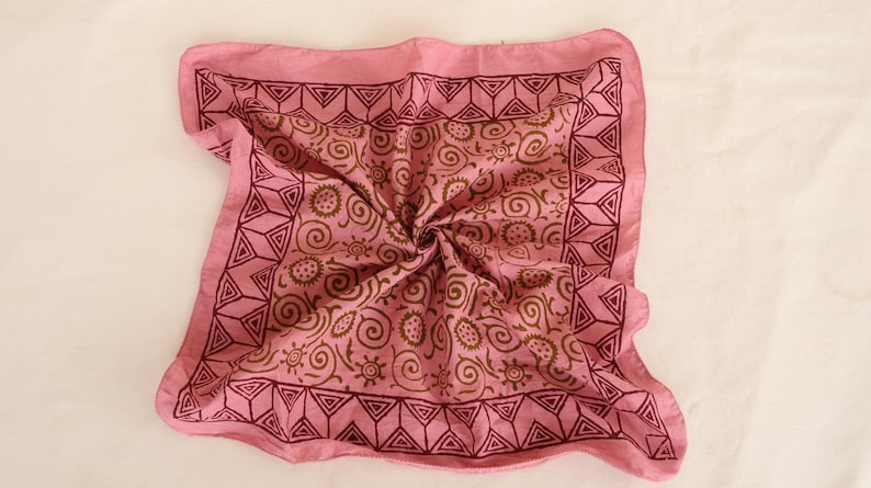 May include: A square pink bandana featuring a dark maroon border with a repeating geometric triangle pattern. The center of the fabric displays a repeating brown print of swirling lines and small sun-like circular motifs across the pink surface.