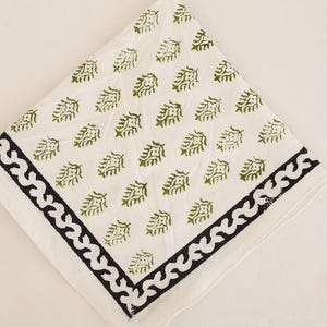 May include: A square white cloth napkin featuring a repeating pattern of small olive green leaf motifs. The napkin is finished with a wide, dark charcoal black border that displays a continuous, stylized wave or scroll design along the edges.