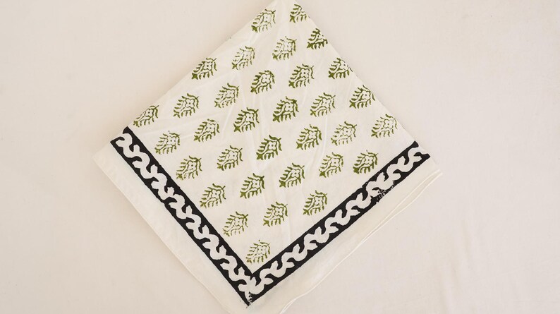 May include: A square white cloth napkin featuring a repeating pattern of small olive green leaf motifs. The napkin is finished with a wide, dark charcoal black border that displays a continuous, stylized wave or scroll design along the edges.