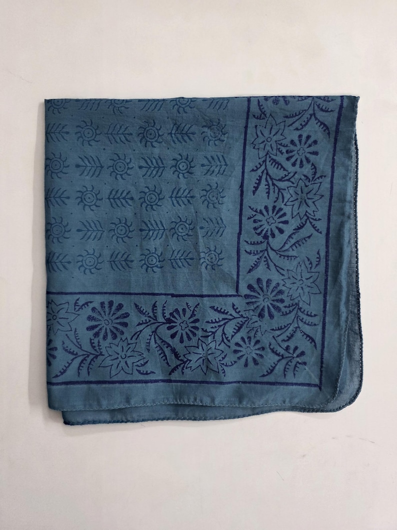 May include: A square blue bandana featuring a dark blue printed pattern. The center displays a grid of small sun-like motifs and dots, while the border is decorated with a repeating design of stylized flowers and leaves on all four sides.