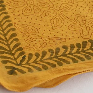 May include: A square fabric textile in a mustard yellow hue featuring a dark olive green leaf-patterned border along the edges and a central design of outlined floral shapes and scattered dots across the main surface of the cloth material.