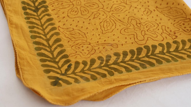 May include: A square fabric textile in a mustard yellow hue featuring a dark olive green leaf-patterned border along the edges and a central design of outlined floral shapes and scattered dots across the main surface of the cloth material.