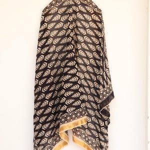 May include: Black and cream-colored scarf with a repeating leaf pattern. The scarf has a gold border with a circular design. The fabric is draped, showcasing the pattern and texture. A stylish accessory.