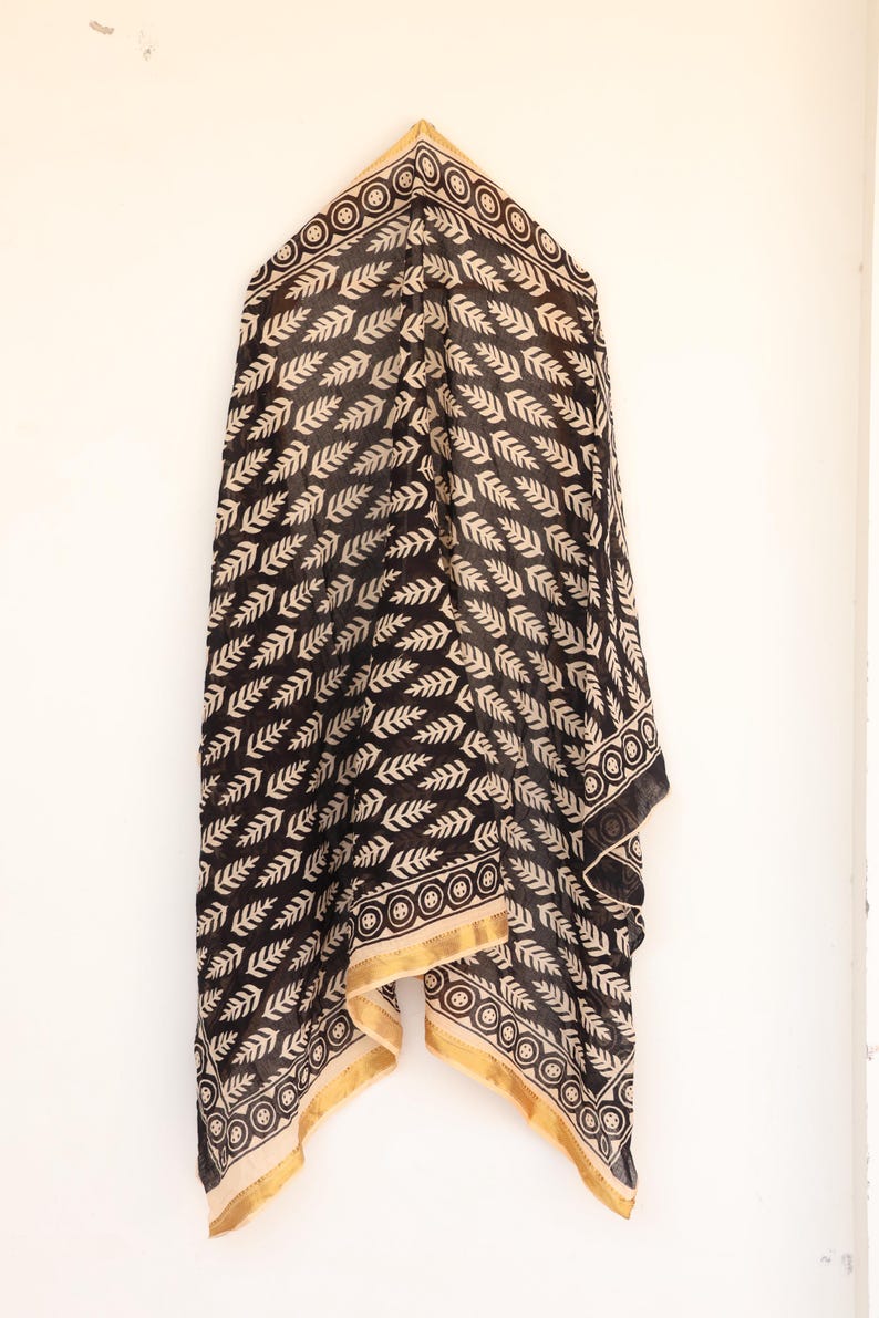 May include: Black and cream-colored scarf with a repeating leaf pattern. The scarf has a gold border with a circular design. The fabric is draped, showcasing the pattern and texture. A stylish accessory.