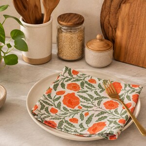 May include: A square cloth napkin featuring a repeating pattern of bright orange poppy flowers and green leafy vines, folded neatly on a round white ceramic plate with a gold-toned metal fork resting on top of the fabric surface.