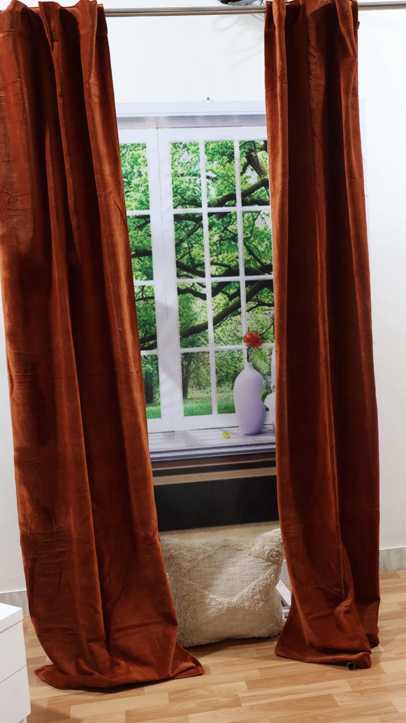 May include: A pair of rust-colored velvet curtains hang from a curtain rod in front of a window with a green tree and a white vase with a red flower.