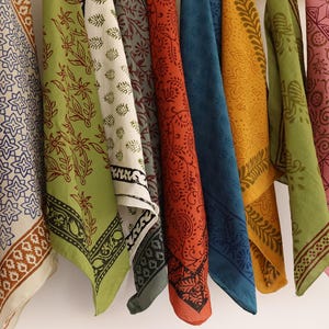 May include: A collection of nine square fabric napkins or handkerchiefs hanging in a row, each featuring distinct block-printed patterns in various colors including maroon, cream, olive green, rust orange, deep blue, mustard yellow, and pink.