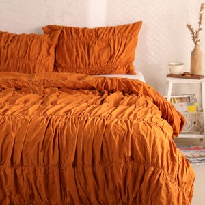 May include: An orange-colored bed set featuring a ruffled design. The set includes a comforter and two pillowcases. The bedding is made of a soft fabric and is displayed on a bed. The overall theme is home decor and bedroom textiles.