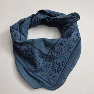 May include: A square bandana in a muted blue shade featuring a darker blue floral print. The fabric displays a repeating pattern of stylized flowers and leaves arranged within a border, folded into a triangular shape with visible hemmed edges.