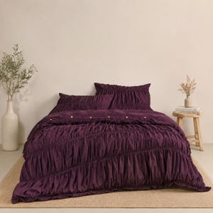 May include: A deep purple bedding set featuring a duvet cover and two matching pillowcases with a gathered, ruched texture. The duvet cover includes a horizontal row of small, light-colored buttons across the top section of the fabric.
