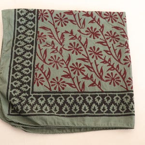 May include: A square fabric scarf in a muted sage green color featuring a dark brown floral pattern of daisy-like flowers on stems. The scarf has a wide border with a repeating dark brown leaf motif along two edges of the folded fabric.