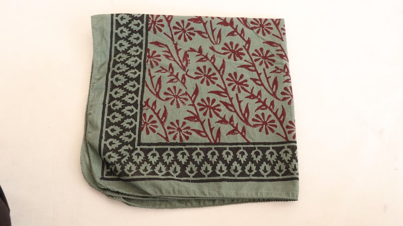 May include: A square fabric scarf in a muted sage green color featuring a dark brown floral pattern of daisy-like flowers on stems. The scarf has a wide border with a repeating dark brown leaf motif along two edges of the folded fabric.