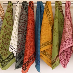 May include: A collection of twelve square fabric napkins hanging from a metal rod. Each napkin features a unique block-printed pattern in various colors including maroon, olive green, mustard yellow, blue, pink, and cream with floral and geometric motifs.