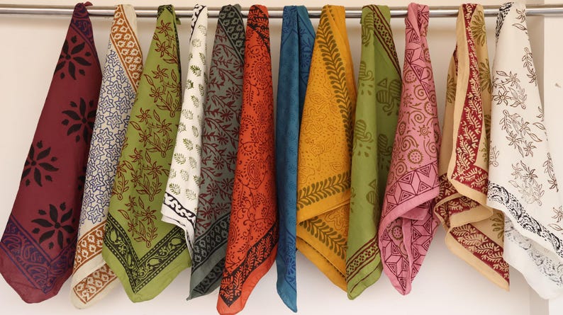 May include: A collection of twelve square fabric napkins hanging from a metal rod. Each napkin features a unique block-printed pattern in various colors including maroon, olive green, mustard yellow, blue, pink, and cream with floral and geometric motifs.