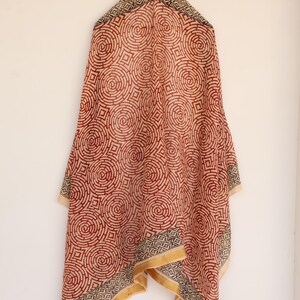 May include: A red and cream-colored scarf with a repeating circular pattern. The scarf has a geometric border in black, cream, and gold. The scarf is hanging against a white wall.