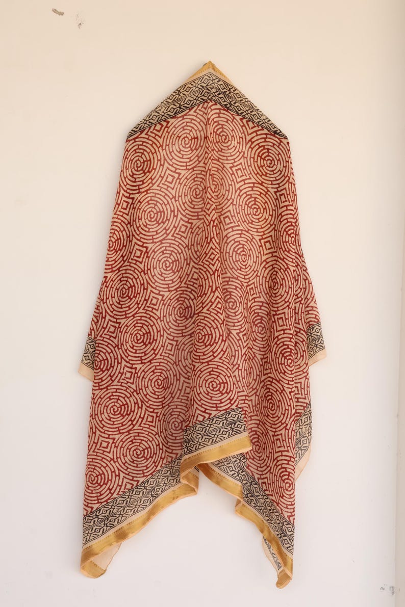 May include: A red and cream-colored scarf with a repeating circular pattern. The scarf has a geometric border in black, cream, and gold. The scarf is hanging against a white wall.