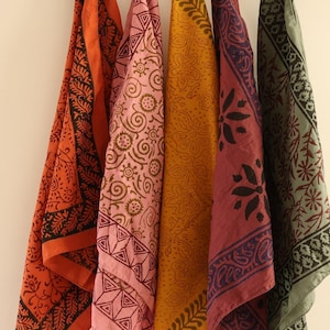 May include: A wooden hanger displays five colorful patterned scarves. The scarves are in shades of orange, pink, yellow, burgundy, and green. Each scarf has a unique black design. The scarves are clipped to the hanger with blue clips.