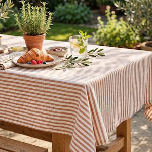 May include: A rectangular tablecloth featuring a pattern of vertical stripes in alternating shades of terracotta orange and white. The fabric drapes over the edges of a wooden table, providing a textured surface for a breakfast setting with a croissant.