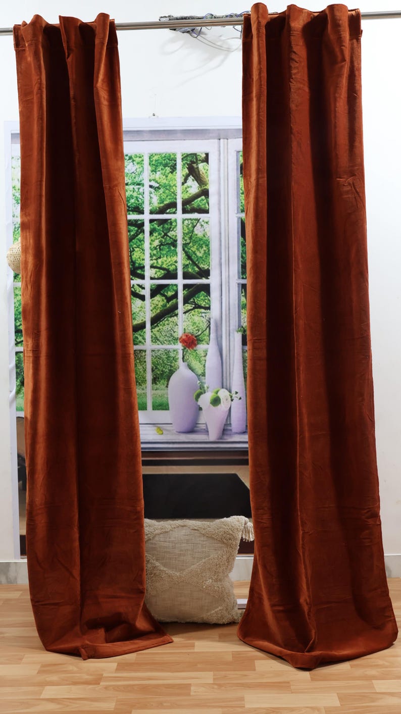 May include: Two rust-colored velvet curtains hang from a curtain rod in front of a window. The curtains are closed and the fabric has a slight sheen.