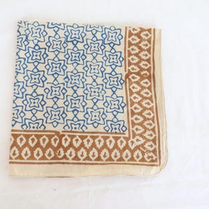 May include: A square textile featuring a cream-colored base with a repeating blue floral pattern across the main section. The border consists of a brown geometric design with leaf-like shapes, framed by a solid cream-colored edge on the perimeter.