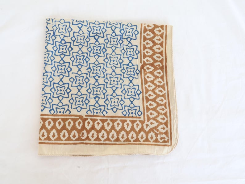 May include: A square textile featuring a cream-colored base with a repeating blue floral pattern across the main section. The border consists of a brown geometric design with leaf-like shapes, framed by a solid cream-colored edge on the perimeter.
