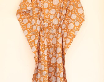 May include: Mustard-colored kaftan dress with a white floral pattern. The dress has a V-neck, a drawstring waist, and wide sleeves. It is hanging on a wooden hanger.