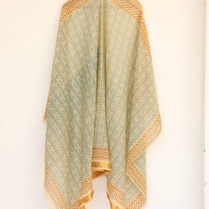 May include: A draped scarf with a geometric pattern in shades of green and beige. The scarf has a gold border and is hanging against a white wall. The fabric appears lightweight and flowing, suitable for warmer weather.