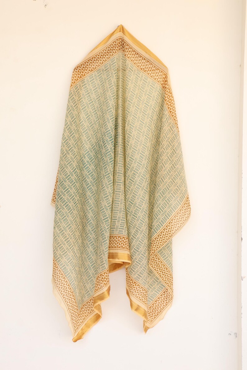 May include: A draped scarf with a geometric pattern in shades of green and beige. The scarf has a gold border and is hanging against a white wall. The fabric appears lightweight and flowing, suitable for warmer weather.