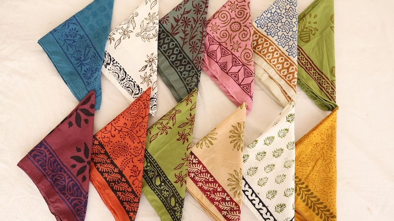 May include: A collection of twelve triangular folded fabric napkins arranged in two rows. Each napkin features a unique block-printed pattern in various colors including blue, white, grey, pink, green, orange, yellow, and maroon with floral motifs.