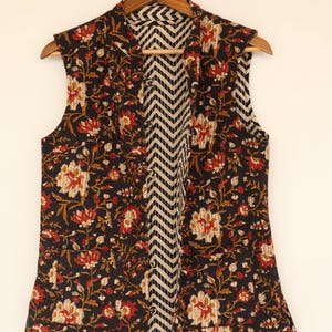 May include: A sleeveless vest with a floral pattern in red, cream, and gold on a black background. The interior lining features a black and white chevron design. The vest is hanging on a wooden hanger.