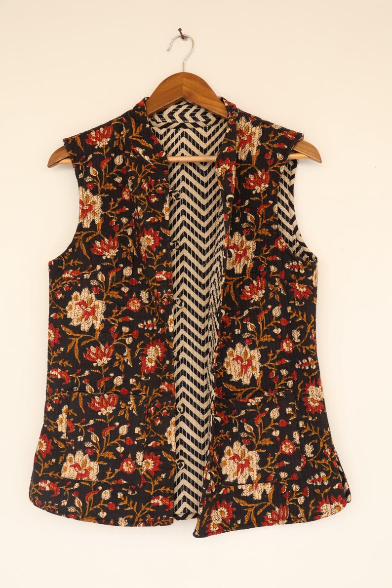 May include: A sleeveless vest with a floral pattern in red, cream, and gold on a black background. The interior lining features a black and white chevron design. The vest is hanging on a wooden hanger.