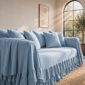 May include: A light blue sofa cover featuring a double-layered ruffled skirt along the base. The cover is draped over the sofa, including the arms, and is accompanied by matching square throw pillows in the same solid light blue fabric.