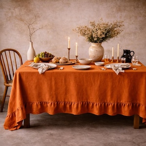 Könnte beinhalten: Ein orangefarbenes Leinentischtuch mit Rüschenkante bedeckt einen Holztisch, der für eine Mahlzeit gedeckt ist. Der Tisch ist mit einer Blumenvase, Kerzen, Tellern und einer Schale mit Trauben geschmückt. Im Hintergrund ist ein Holzstuhl zu sehen.