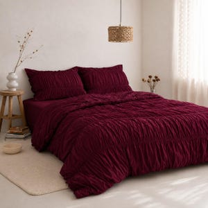 May include: A deep burgundy bedding set featuring a comforter and two matching pillow shams with a gathered, textured ruched design. The bedding covers a bed frame, creating a uniform dark red appearance across the surface of the mattress.