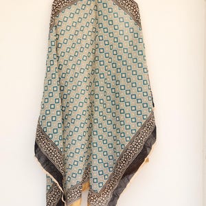 May include: A patterned scarf with a geometric design in teal, beige, and black. The scarf has a square pattern with a decorative border and gold trim. The scarf is hanging against a white background.
