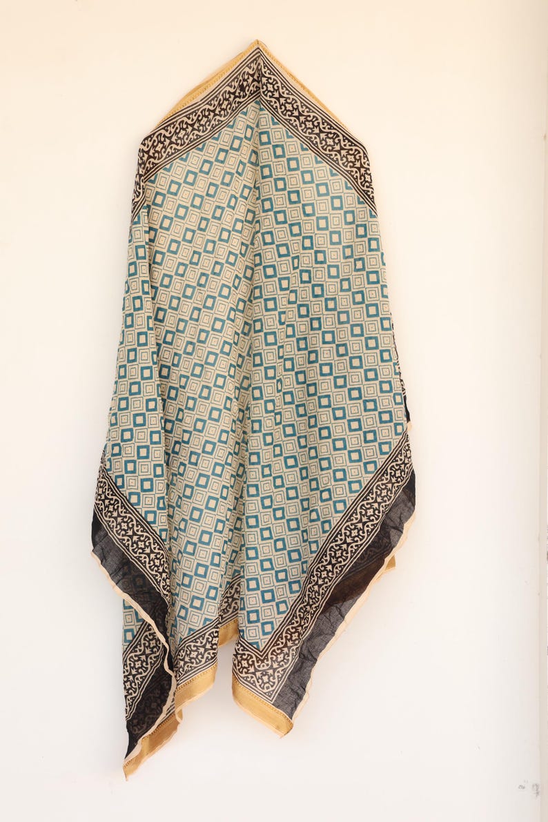 May include: A patterned scarf with a geometric design in teal, beige, and black. The scarf has a square pattern with a decorative border and gold trim. The scarf is hanging against a white background.