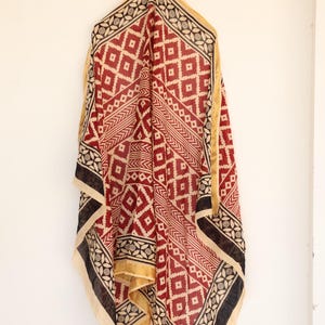 May include: A draped textile with a geometric pattern in red, black, and cream. The scarf features diamond and linear designs, with a gold border. The fabric is hanging against a white wall.