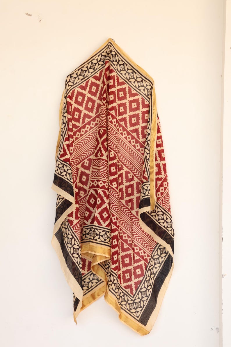 May include: A draped textile with a geometric pattern in red, black, and cream. The scarf features diamond and linear designs, with a gold border. The fabric is hanging against a white wall.
