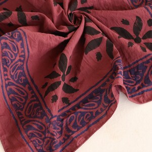 May include: A square fabric bandana in a deep maroon red color featuring a dark navy blue border with a repeating paisley pattern and a central floral motif. The fabric is gathered in the center, showing a soft texture and hemmed edges.