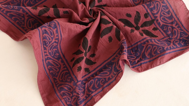 May include: A square fabric bandana in a deep maroon red color featuring a dark navy blue border with a repeating paisley pattern and a central floral motif. The fabric is gathered in the center, showing a soft texture and hemmed edges.
