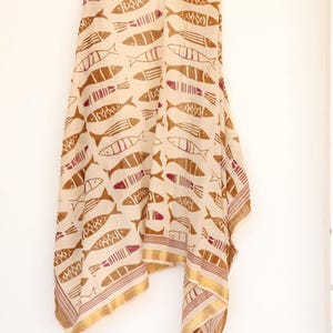 May include: A beige scarf with a repeating pattern of brown fish illustrations. The scarf has a gold border with a thin maroon stripe. The fabric is draped, showcasing the design and texture.